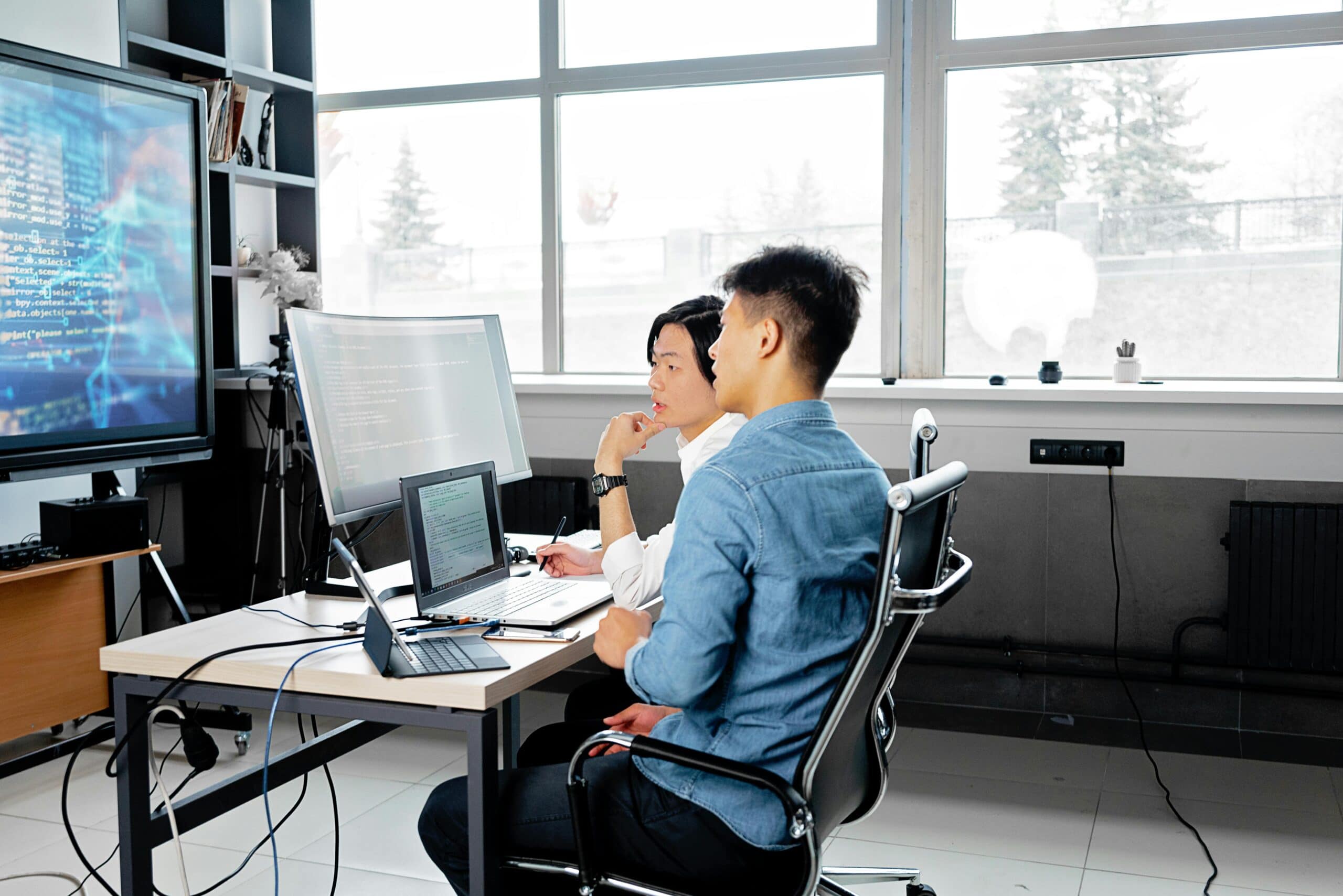 pexels-mikhail-nilov-7988748 (1) Two male workers sitting next to eachother at a desk with paperwork and tech devices while looking at a large computer screen together.