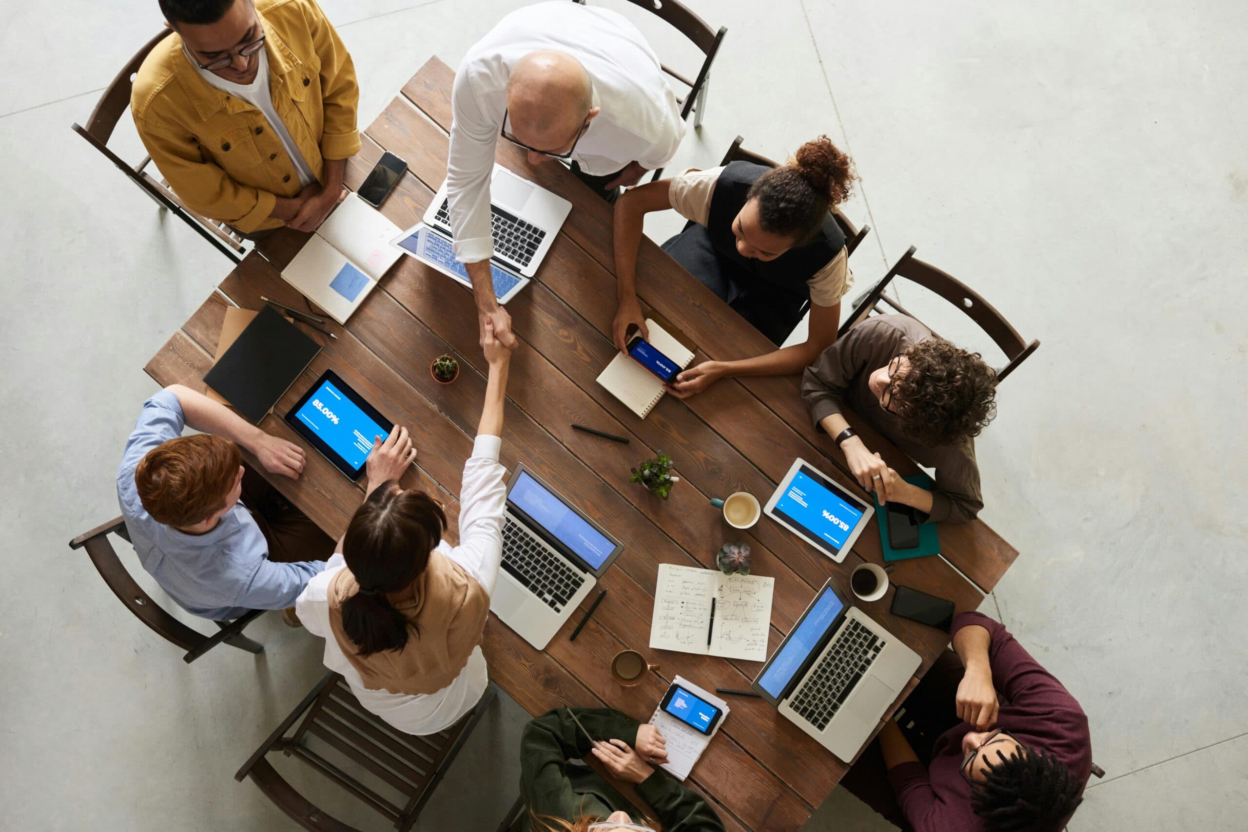 pexels-fauxels-3183197 (1) A team of professionals sitting around a large table with laptops and other technology, shaking hands and conversing.